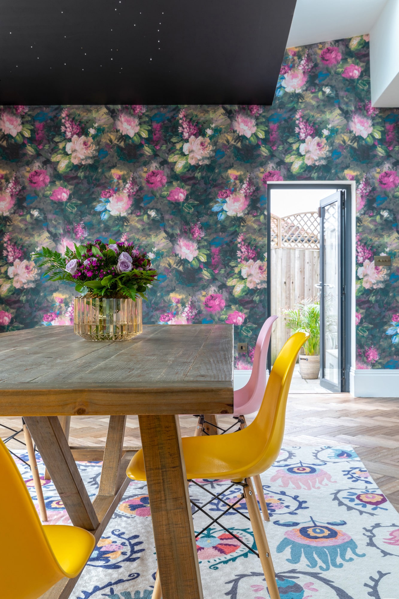 Interior design photo: dining area with floral wallpaper, wooden table, plastic yellow and pink chairs; black ceiling