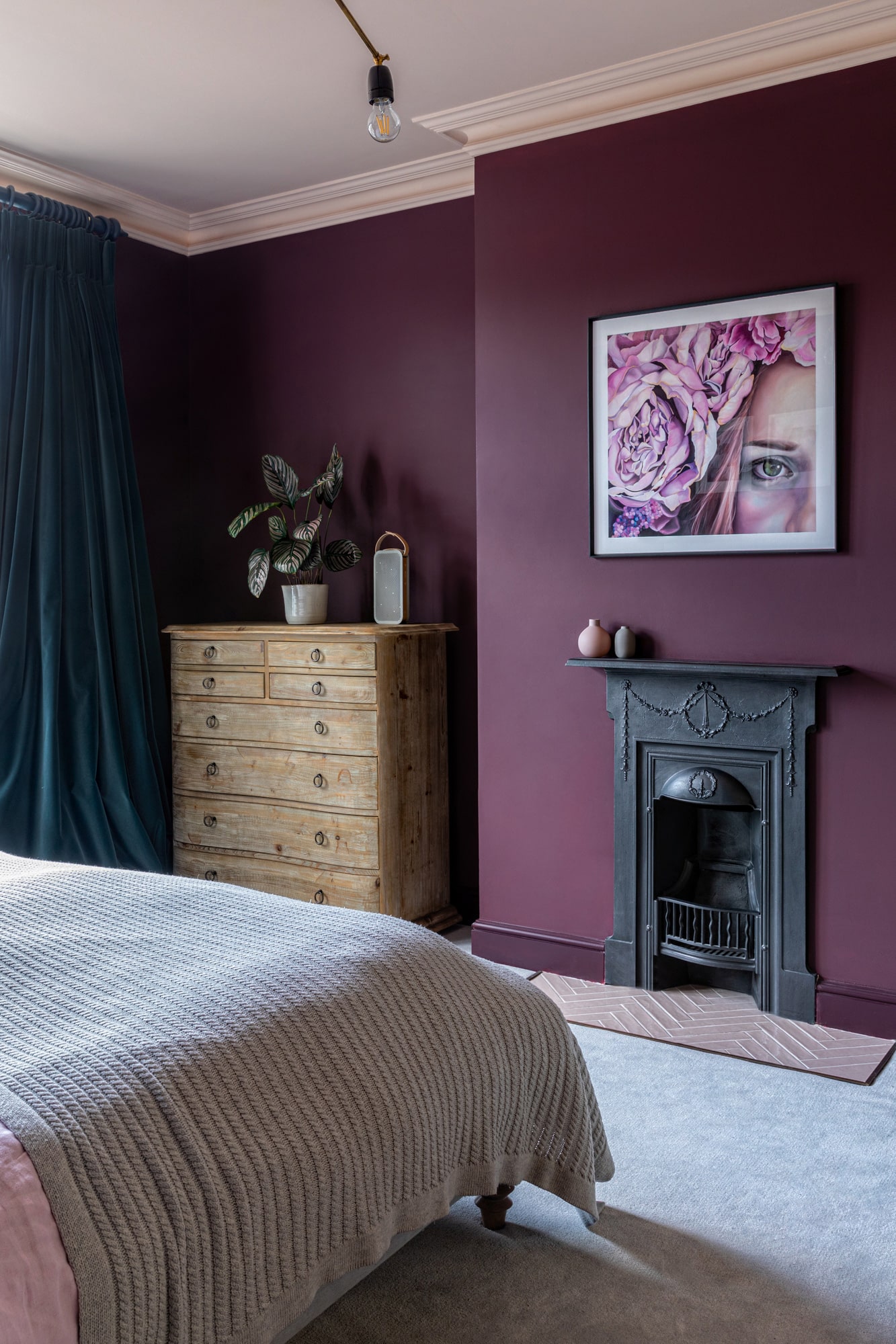 Interior design photography: bedroom with dark burgundy walls;  art with a girl in a flower band; fireplace; teal velvet curtains; bed; wooden chest of drawers