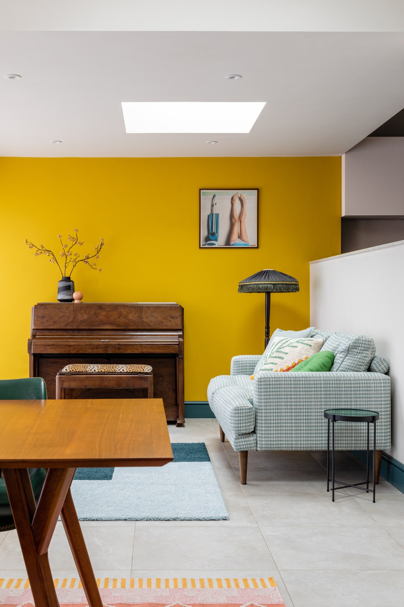 Interior design photo: a view from a dining area to a sitting living room area with a wooden table, a yellow wall, a brown piano and a sofa upholstered with green gingham pattern