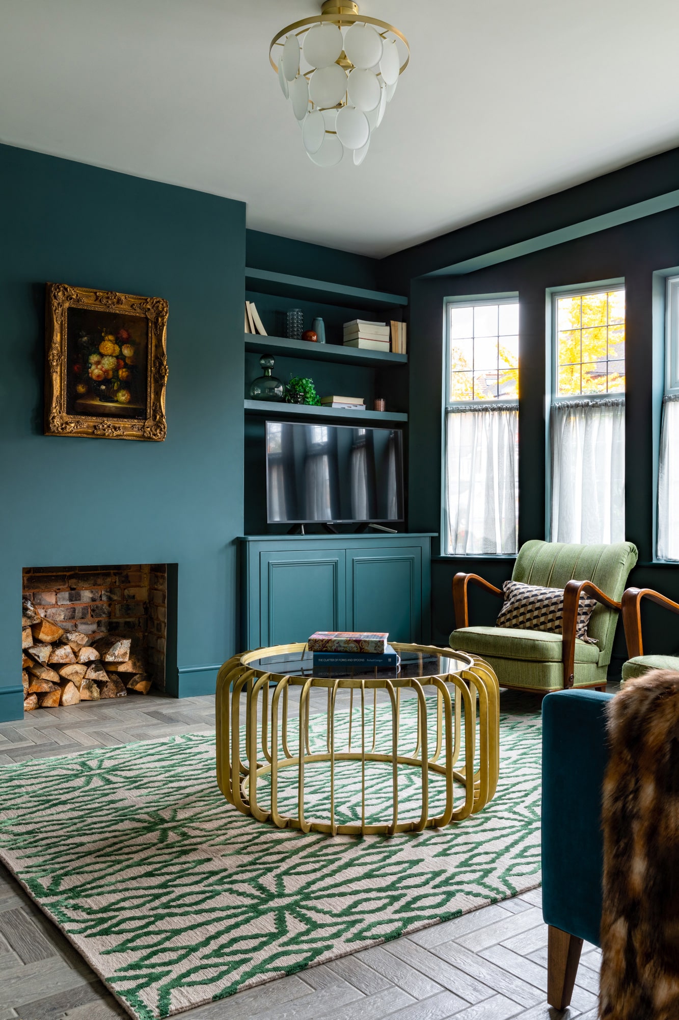  interior design photography: a sitting room with dark blue walls, a fireplace with wood logs, built in shelves and cardboard with a tv and books. There 2 green armchairs next to the window and a gold metal coffee table with a glass top in the middle of the room.