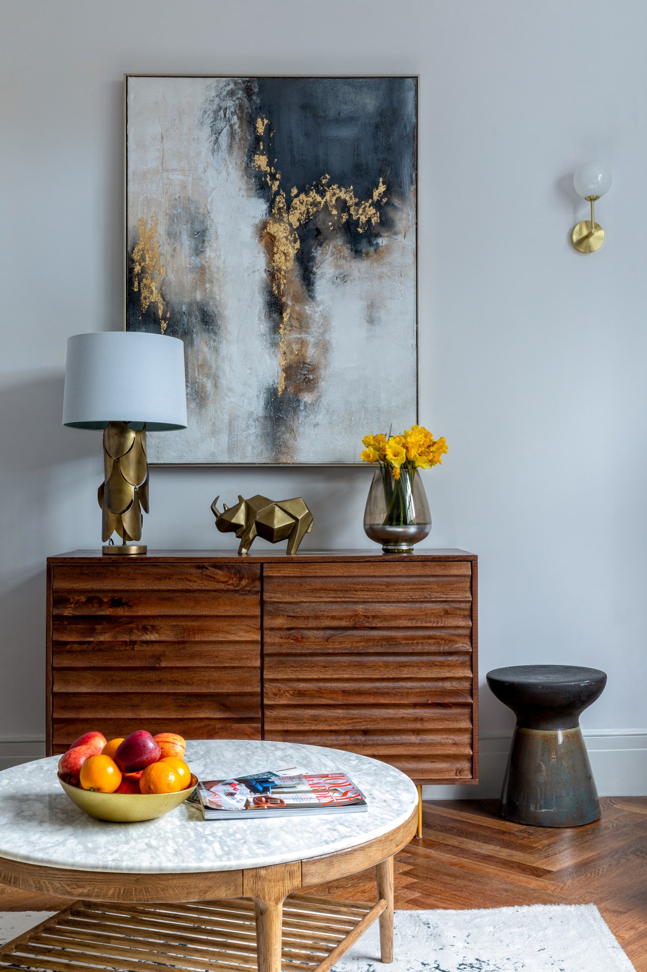 interior detail shot: living room with blue walls, wooden cabinet with lamp and accessories on top; wooden  coffee table with marble top, fruit vase and magazines on it; abstract painting on the wall