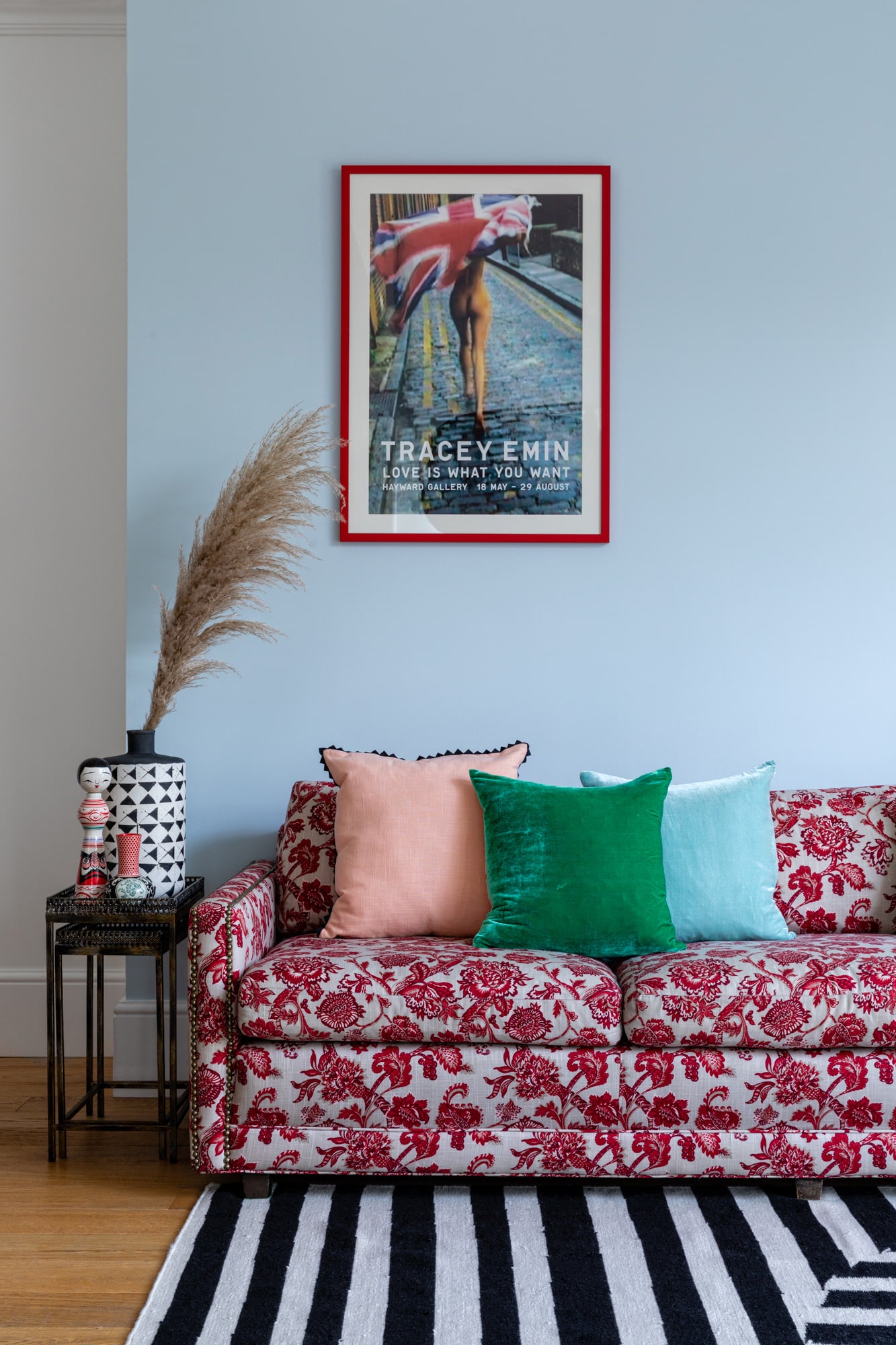 Interior photo of a living room: light blue walls; sofa with red pattern; colourful cushions; Tracey Emin poster; black and white striped rug