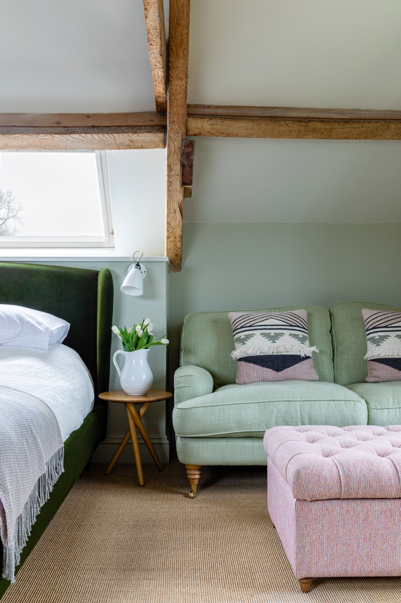 renovated barn bedroom: olive green walls; dark green velvet bed with white linen; bedside table with a vase of tulips; light green sofa and pink soft stool