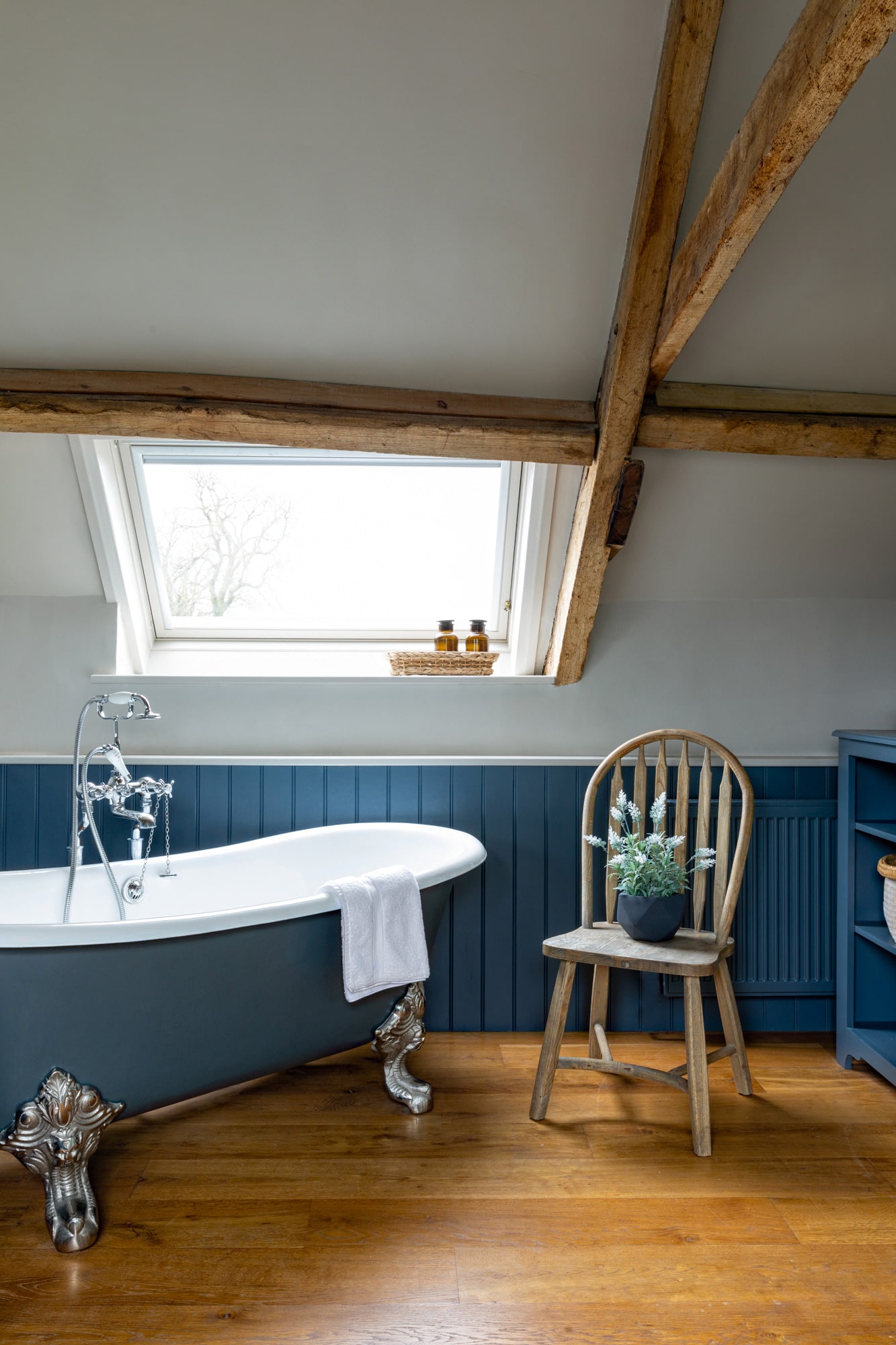 photo of a barn bathroom with freestanding bath; window; blue paneling; a chair with a plant pot on it