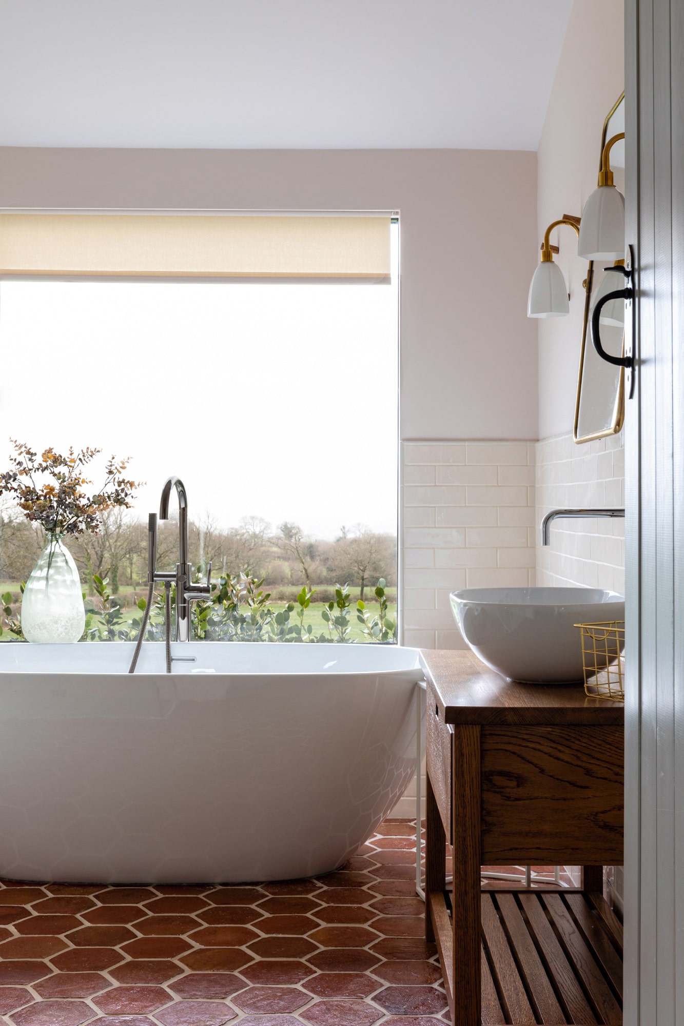 interior photo of a bathroom: round freestanding bath in front of a window with a field view; a round sink with a wooden cupboard